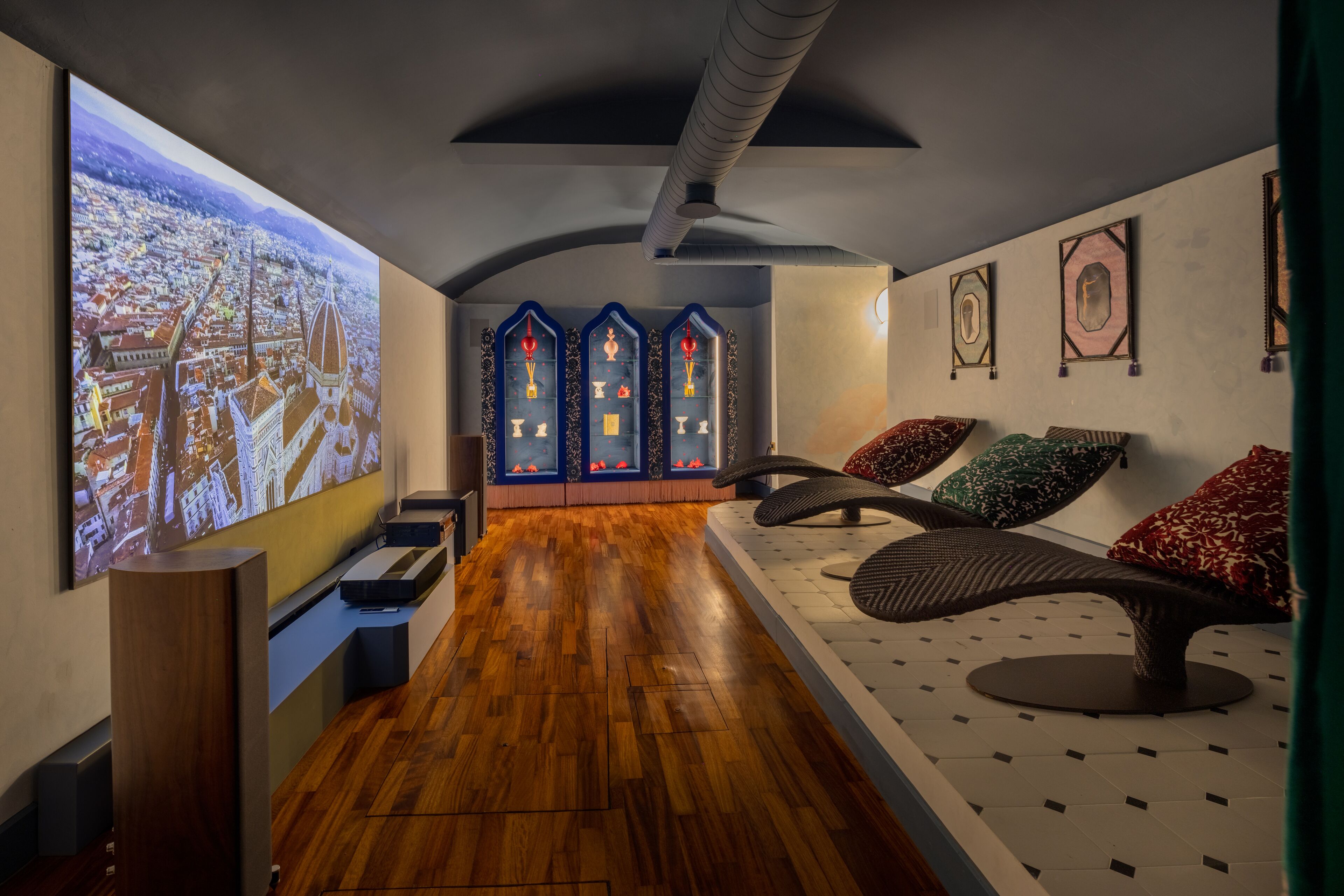 Una camera d'albergo moderna con pavimento in legno, un grande schermo di proiezione che mostra una vista sulla città, una zona salotto originale con cuscini colorati e un mobile decorativo con ante in vetro.