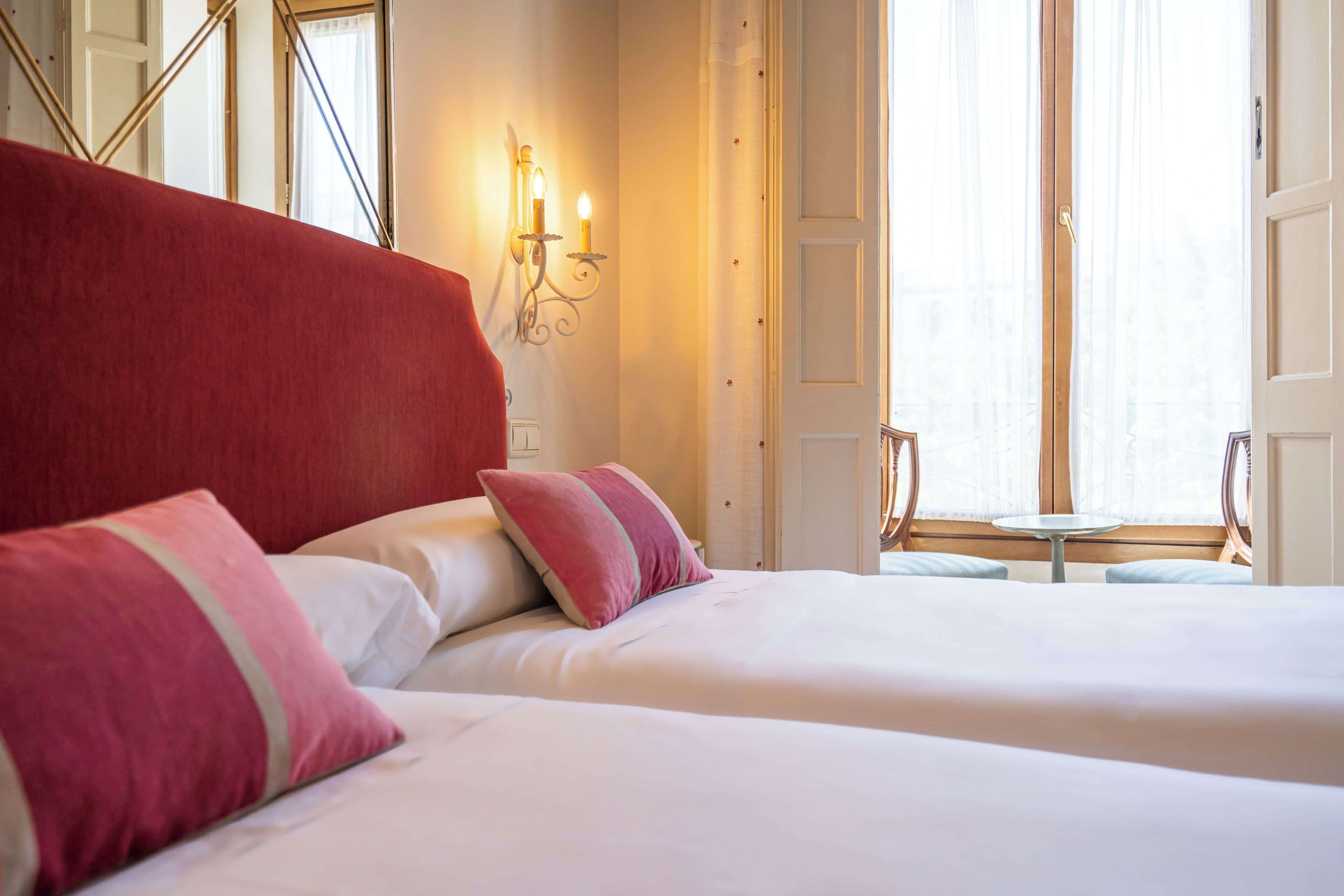 Une chambre d’hôtel avec deux lits, une tête de lit rouge et une petite table près de la fenêtre.