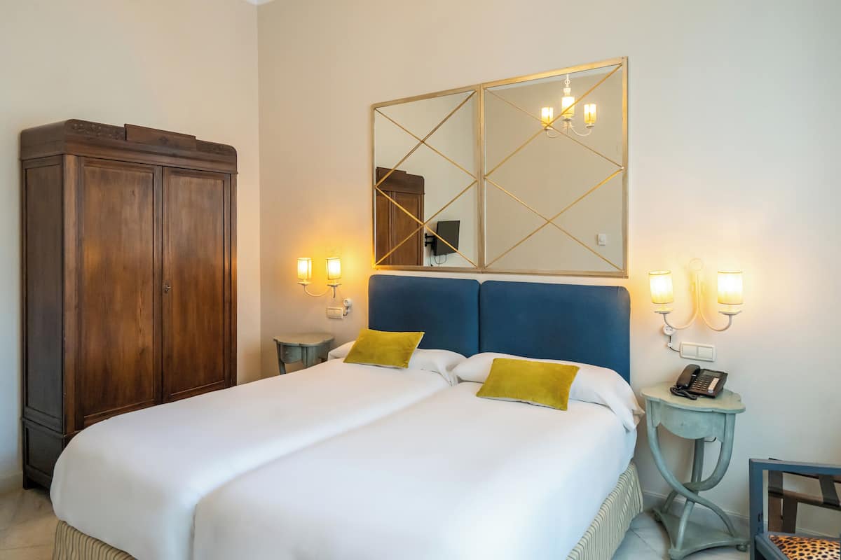 A neatly made bed with white linens and two mustard pillows, a wooden wardrobe, a bedside table with a phone, and wall-mounted lights.