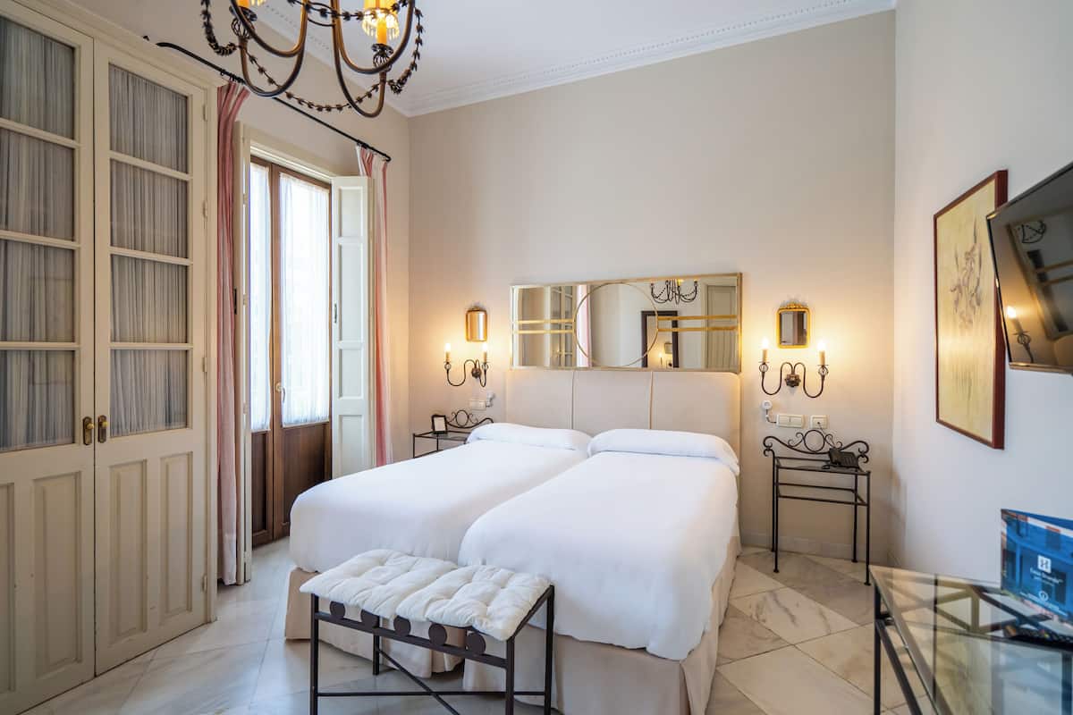 A bedroom with two beds, a large mirror, bedside lamps, a bench, and a glass table.