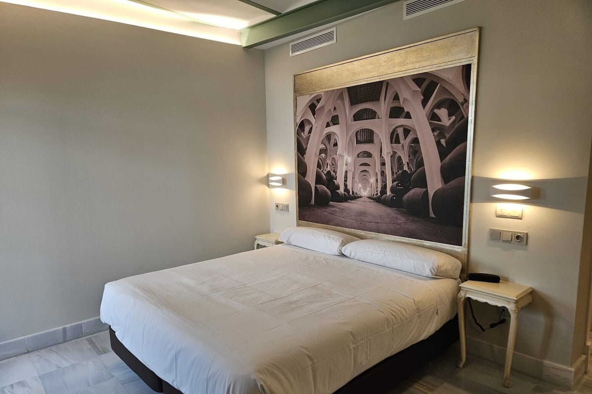 A neatly made bed in a hotel room with a large framed picture on the wall, two bedside tables, and a light switch.