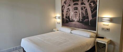 A neatly made bed in a hotel room with a large framed picture on the wall, two bedside tables, and a light switch.