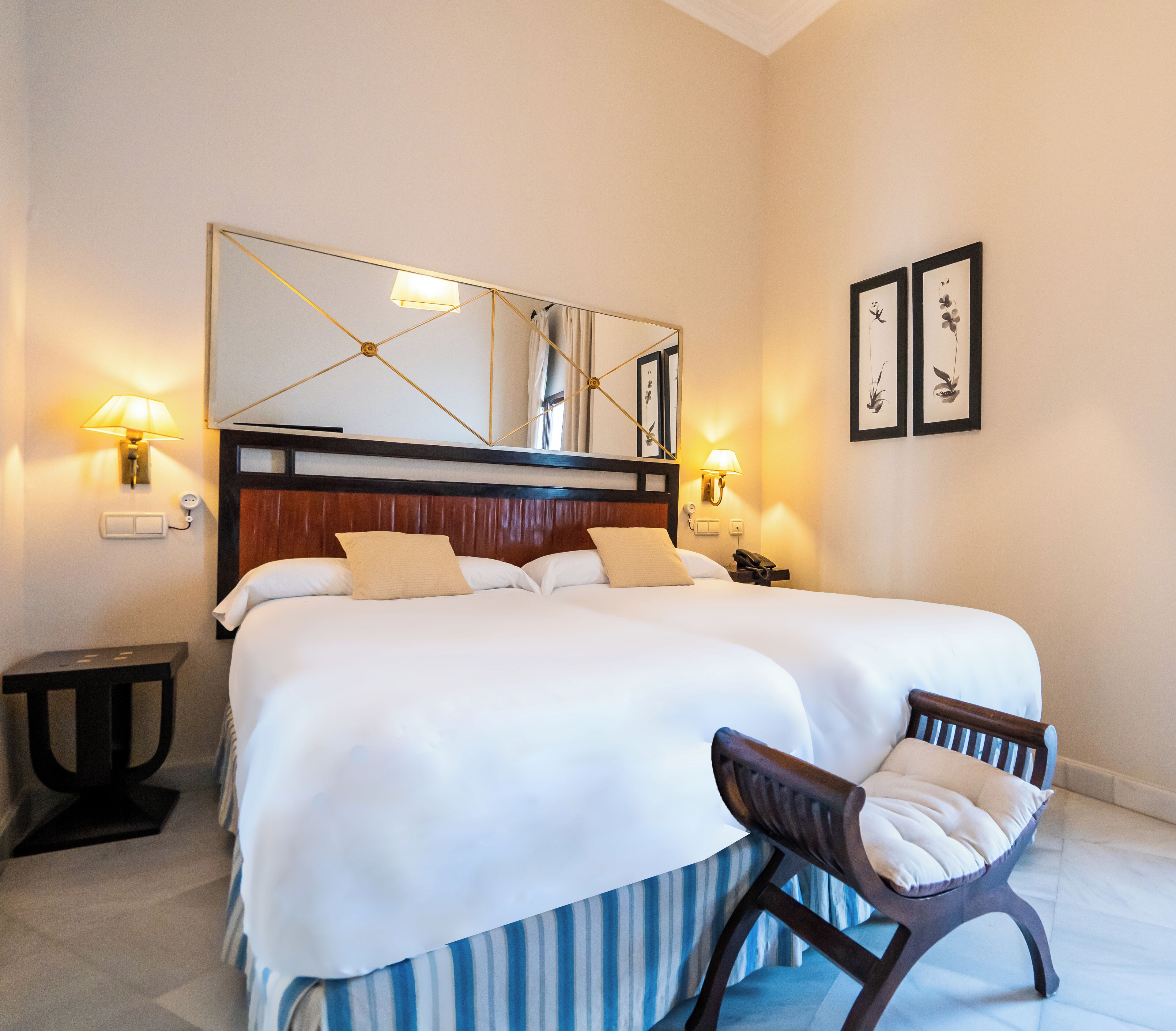 Une chambre d’hôtel avec un grand lit, deux lampes de chevet, une table de chevet, une chaise, un miroir et des œuvres d’art encadrées accrochées au mur.