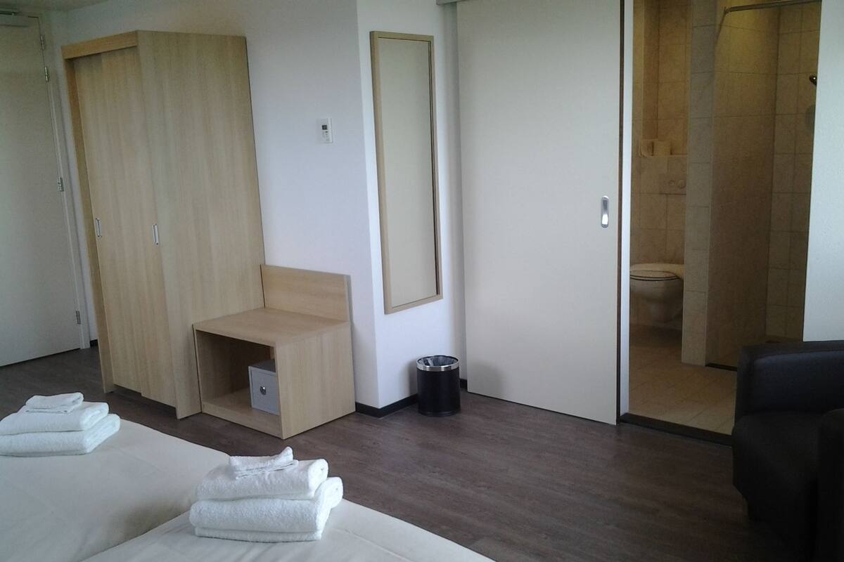 A hotel room with a bed, a wooden wardrobe, a small bench, a trash bin, and a bathroom visible through an open door.