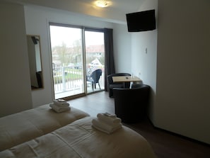 Ein Hotelzimmer mit zwei Betten, einem Schreibtisch, einem Sessel, einem Fernseher und einem Balkon mit Blick auf Gebäude und Bäume.