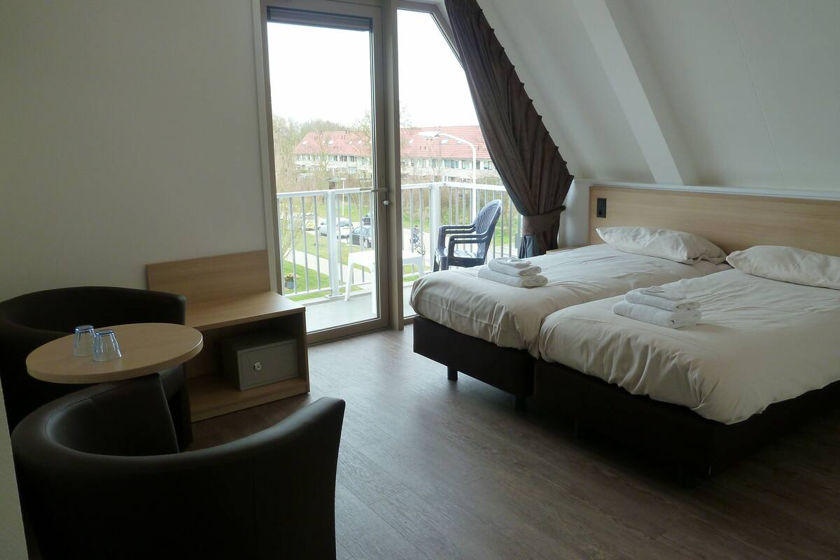A hotel room with a bed, a chair, a small table, and a balcony with a view of buildings.