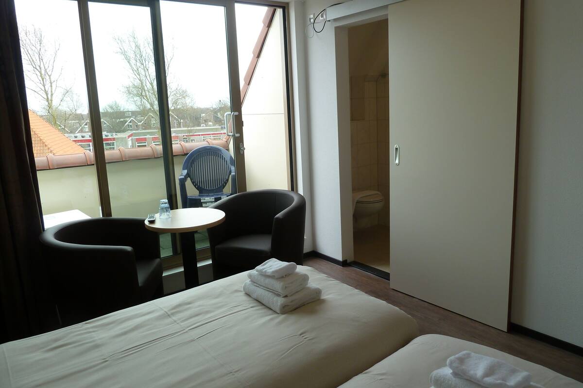 A hotel room with a bed, a small table with a chair, a view of the outside through large windows, and a closet.