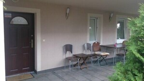 Uma área de estar coberta ao ar livre, com porta de madeira, cadeiras de metal e uma pequena mesa.