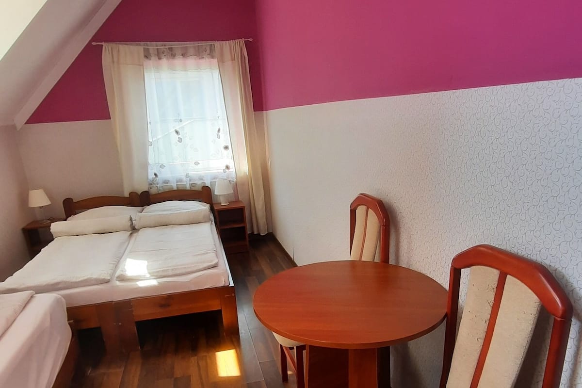Ein Zimmer mit Holzboden, einem runden Tisch, zwei Stühlen, einem Bett und einem Fenster mit Vorhängen.