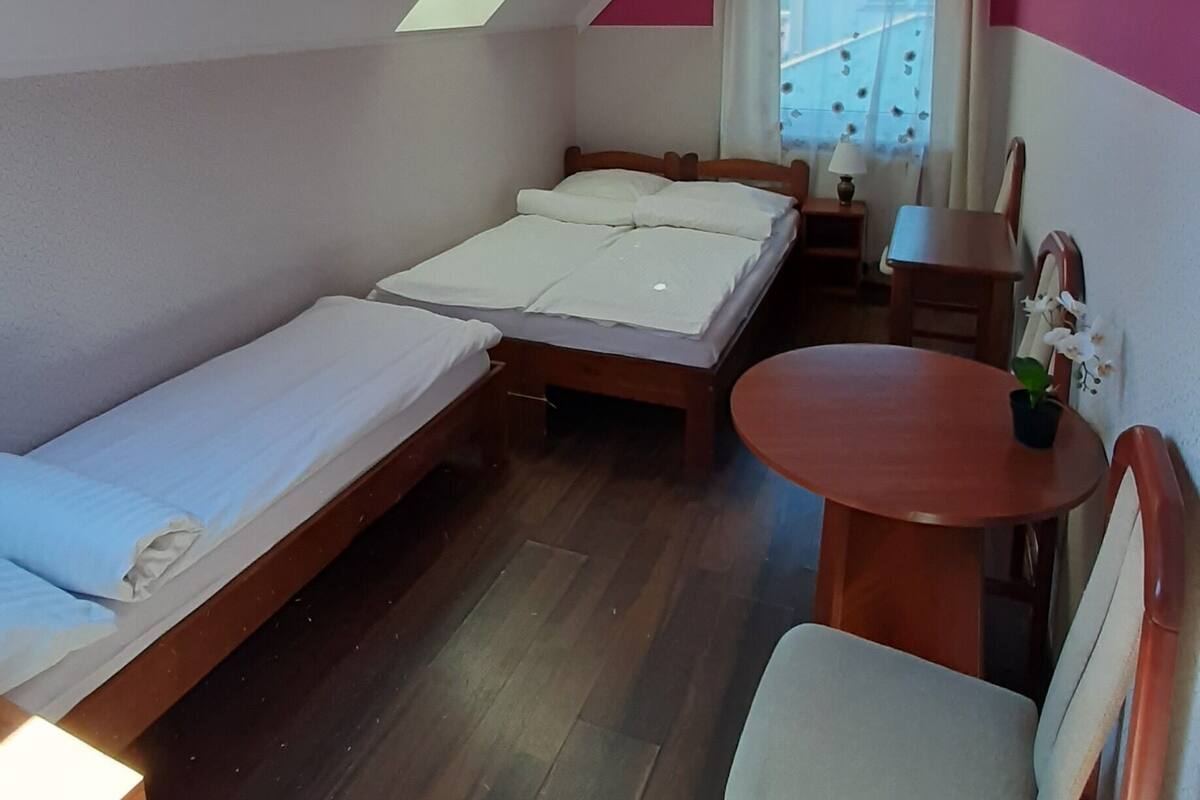 Ein kleines Zimmer mit einem Bett, einem Tisch und einem Stuhl. Über dem Bett ist ein Oberlicht und ein Fenster mit Vorhängen.