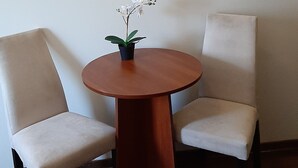 Um quarto pequeno com uma mesa redonda de madeira, duas cadeiras brancas, uma orquídea em vaso e um espelho com moldura de madeira na parede.