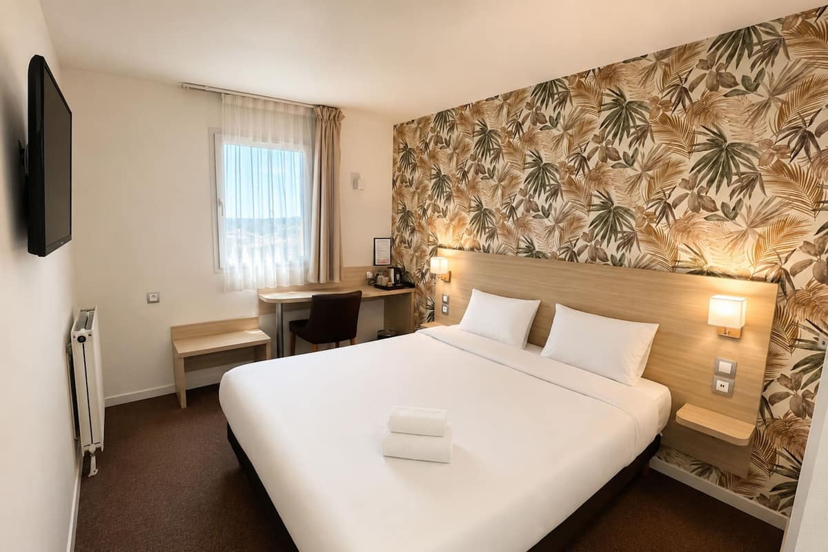Habitación de hotel con una cama grande, un escritorio, una silla, un televisor y una pared con diseño de hojas tropicales.