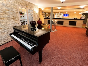 A grand piano in a lobby with a stone wall, a reception desk, and a bar area. - Hôtel Altina (Pacy-sur-Eure)