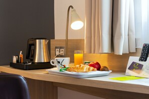 Un petit-déjeuner servi en chambre d’hôtel, comprenant une machine à café, une assiette de croissants, des fruits, un verre de jus d’orange et une lampe.