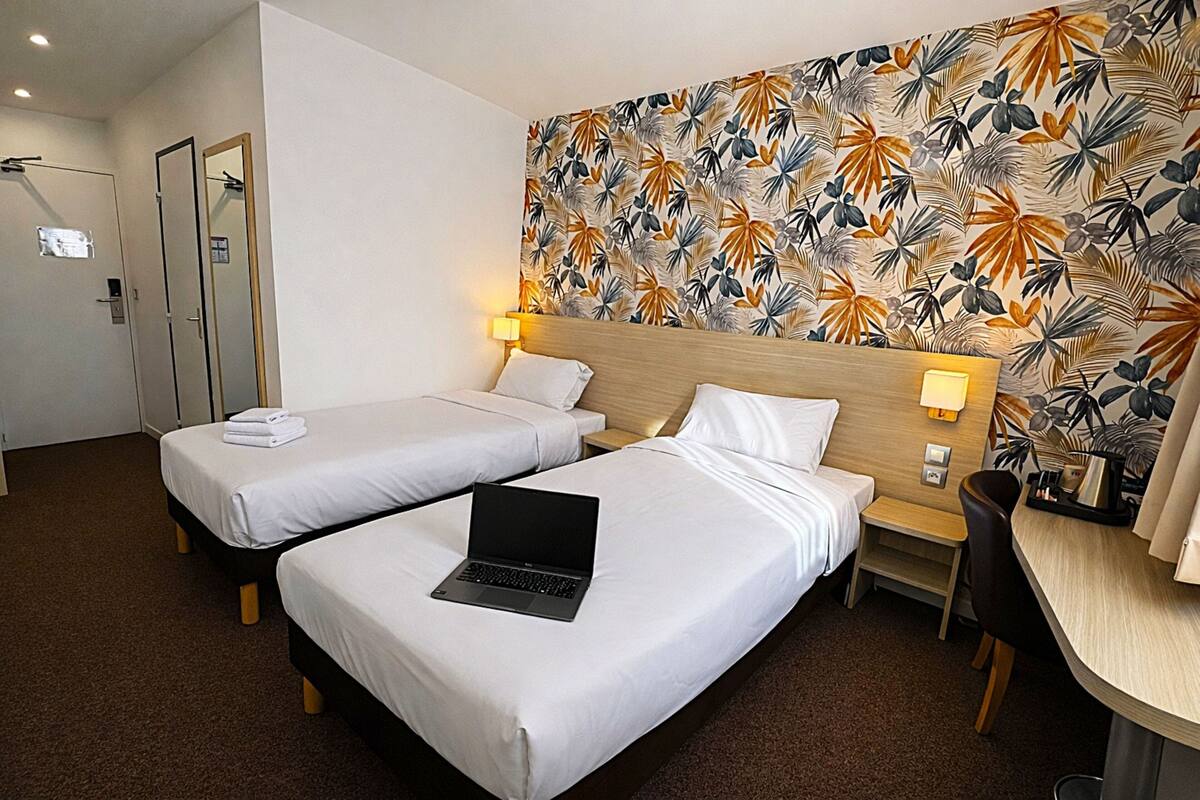 Habitación de hotel con dos camas individuales, un escritorio, un ordenador portátil sobre una cama y una pared con diseño de hojas tropicales.