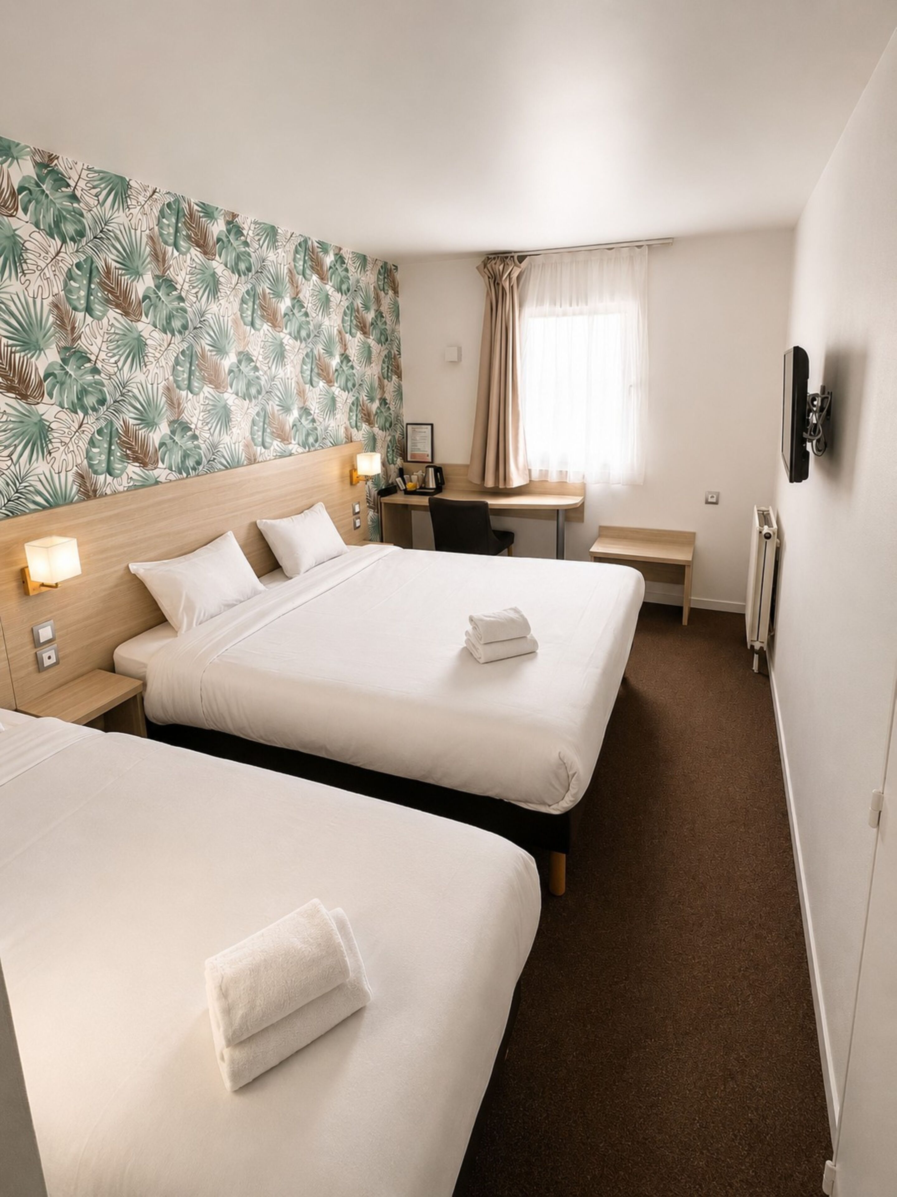 Ein Hotelzimmer mit zwei Betten, einem Schreibtisch, einem Stuhl, einem kleinen Tisch, einem Heizkörper und einer Wand mit einem tropischen Blattmuster.