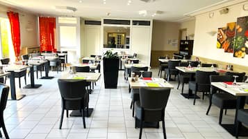 Un restaurante con varias mesas y sillas, una jardinera negra en el centro y una zona de cocina al fondo.