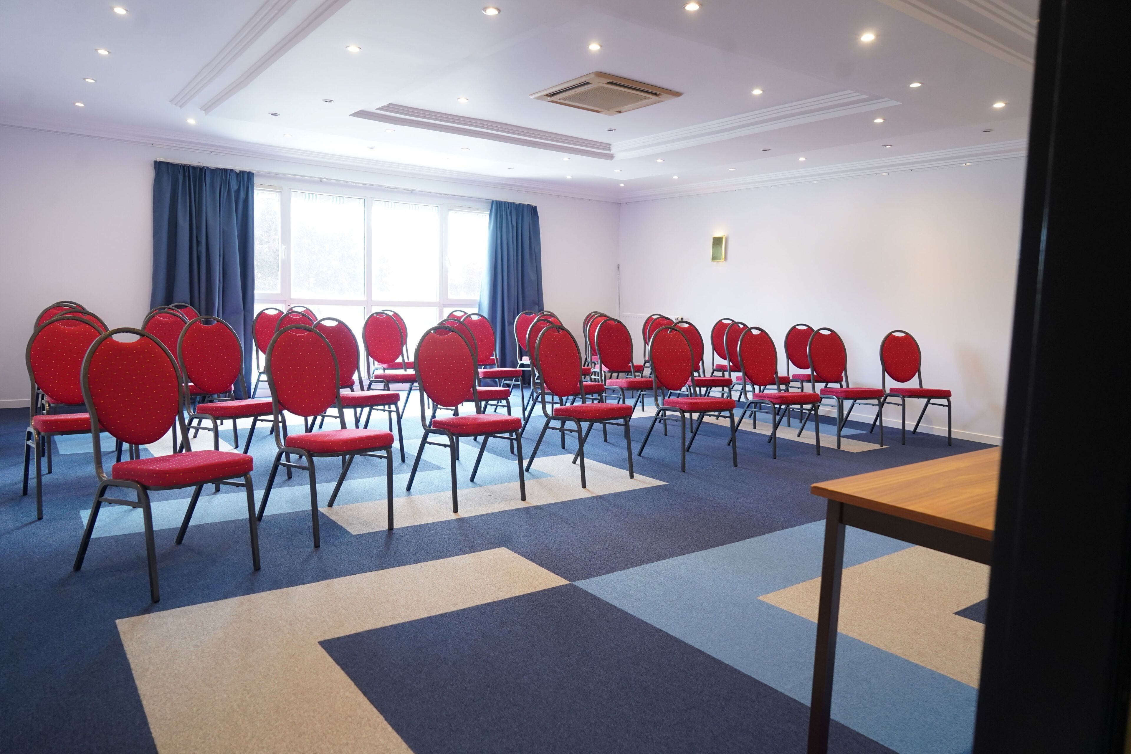 Un salón de conferencias con filas de sillas rojas, una mesa de madera y una alfombra estampada.