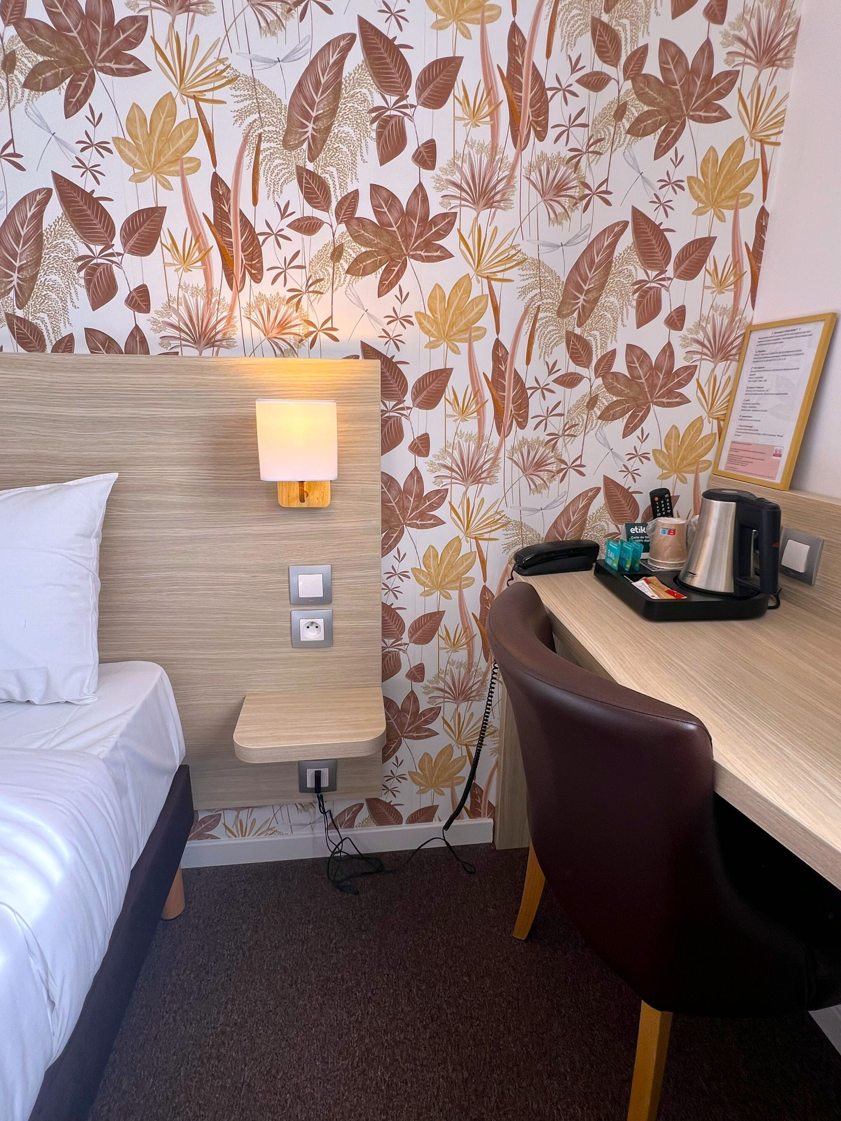 A hotel room with a bed, a desk with a lamp, a chair, a coffee maker, and a wall with a floral wallpaper.