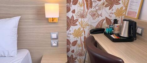 Quarto de hotel com cama, escrivaninha com luminária, cadeira, cafeteira e parede com papel de parede floral.