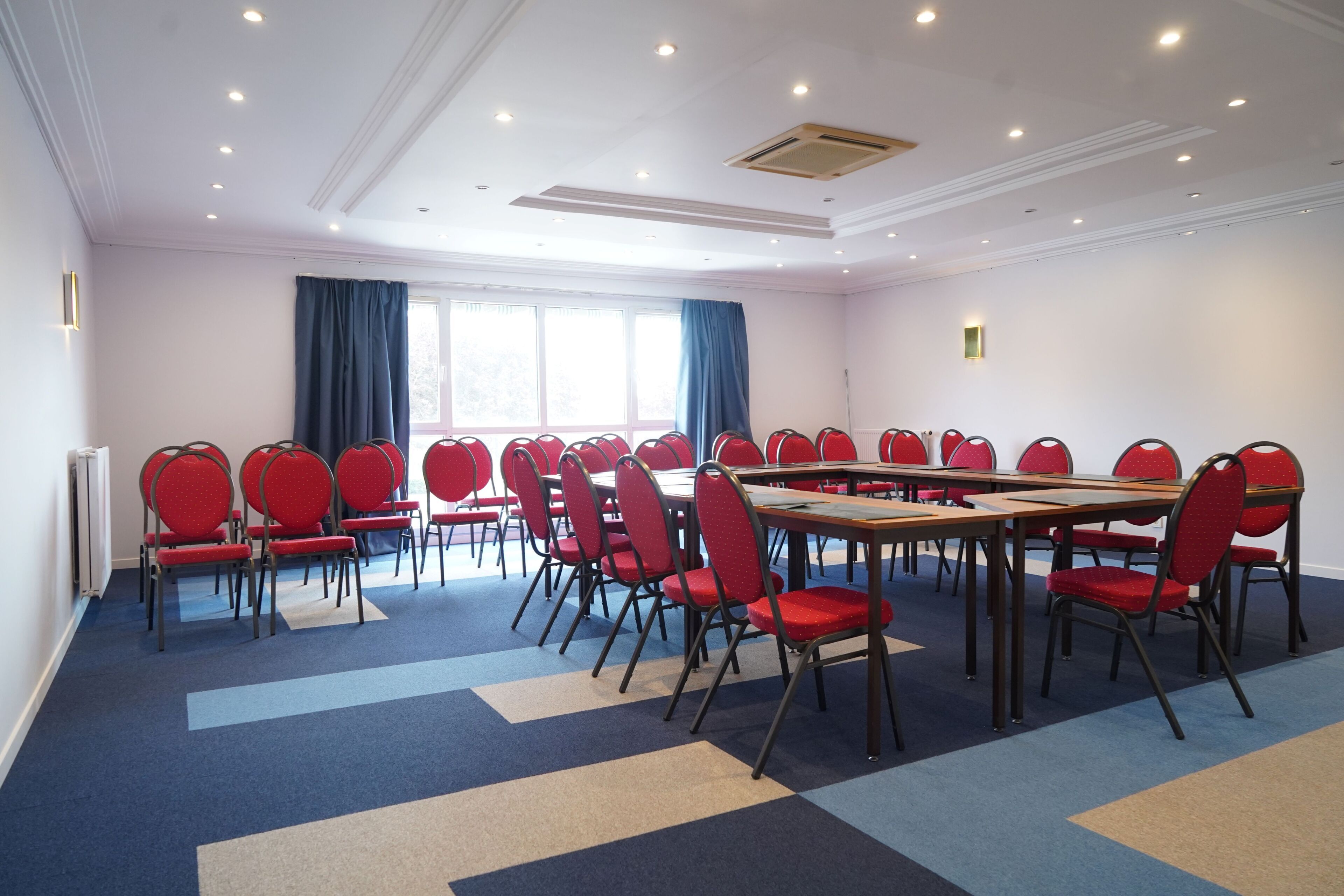 A conference room with multiple tables and chairs, large windows with curtains, and a carpeted floor.
