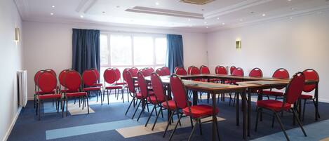 A conference room with multiple tables and chairs, large windows with curtains, and a carpeted floor.