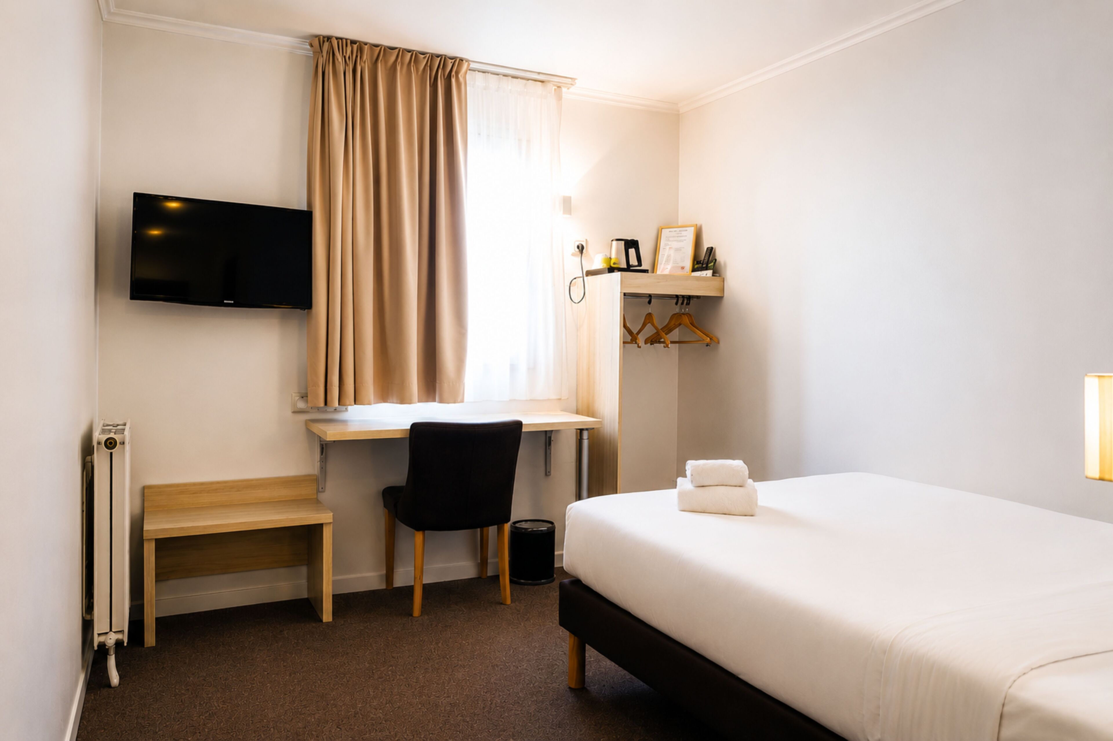 A hotel room with a bed, a desk with a chair, a television mounted on the wall, and a window with curtains.
