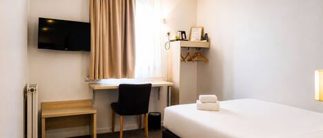 Una habitación de hotel con cama, escritorio con silla, televisor montado en la pared y ventana con cortinas.