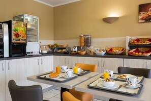 A breakfast setup with a coffee machine, fruit baskets, and breakfast items on trays. - Hôtel Altina (Pacy-sur-Eure)
