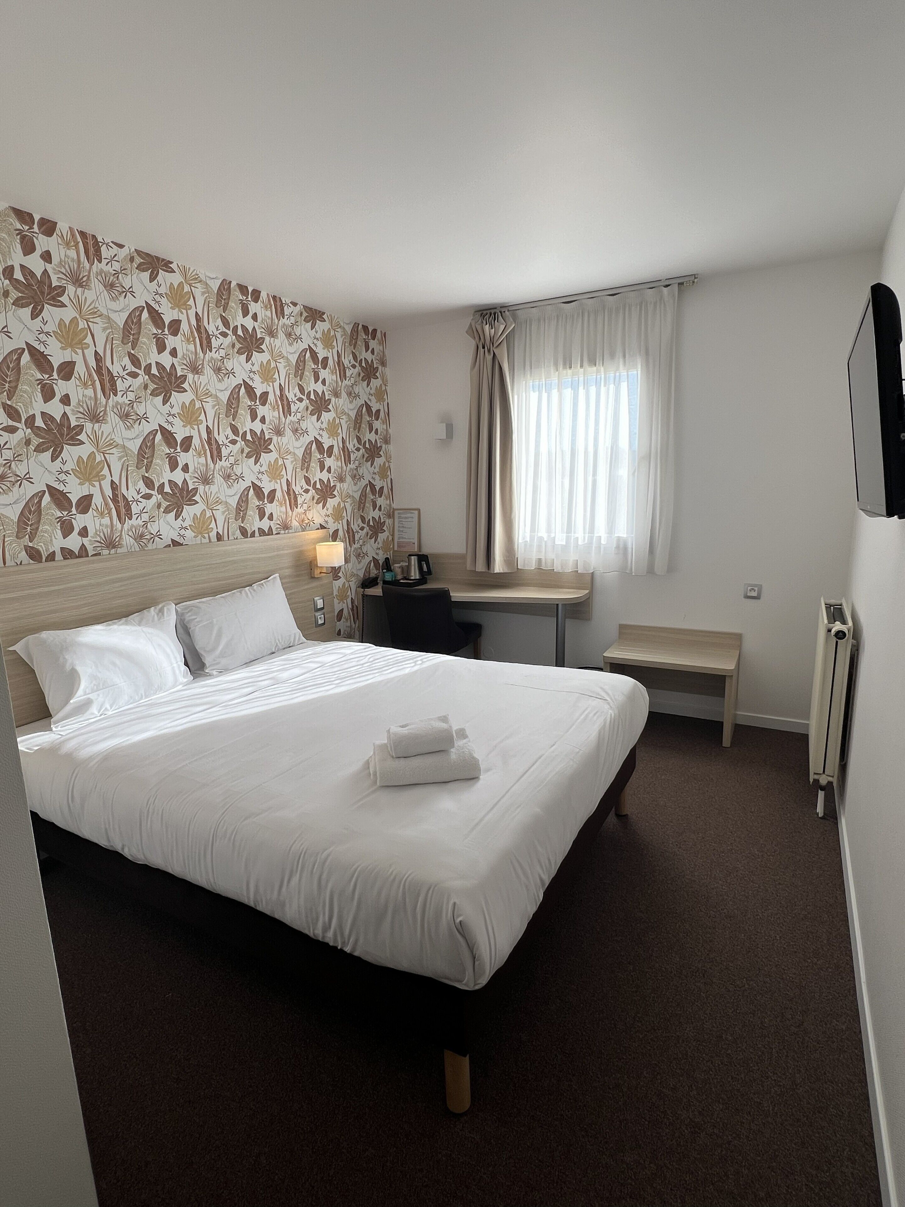 Habitación de hotel con cama, escritorio, silla, mesita de noche, espejo y ventana con cortinas.