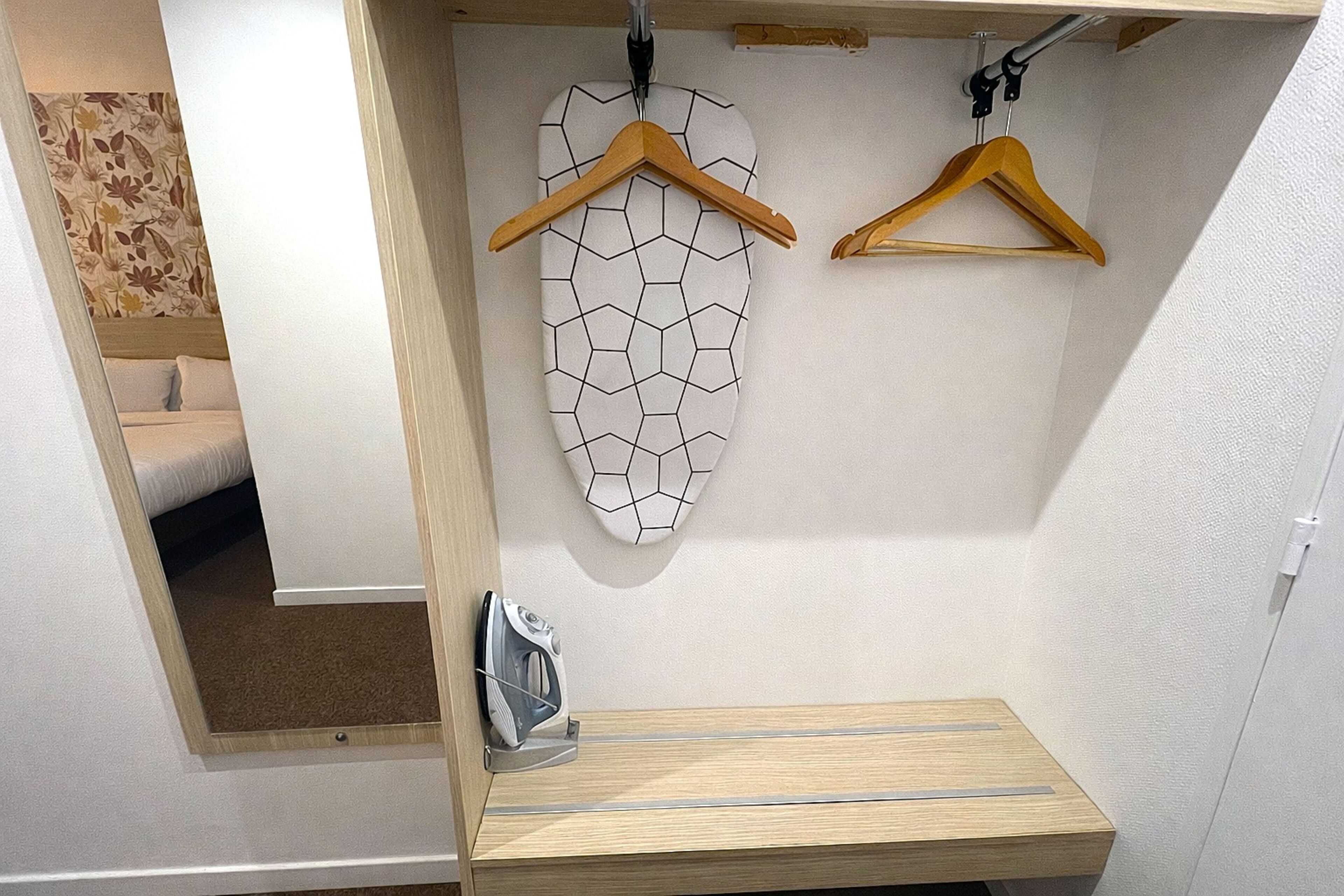 A hotel room with a bed, a wooden shelf, an ironing board, and two wooden hangers.