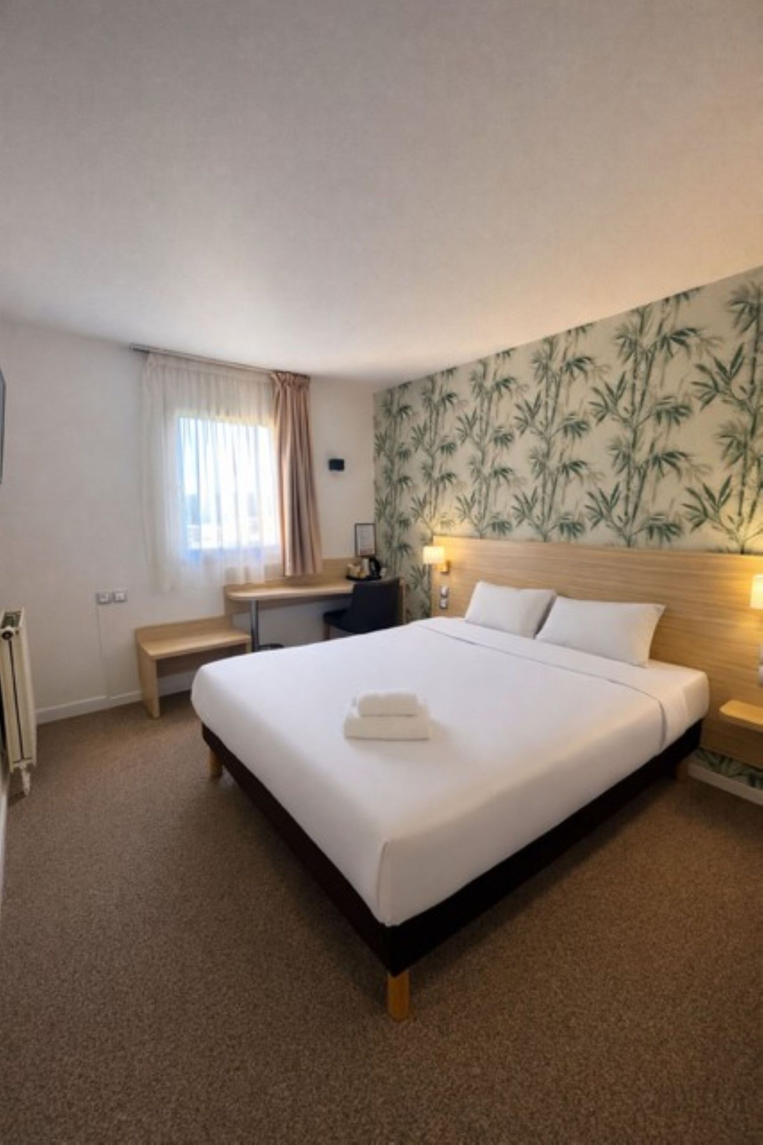 A hotel room with a large bed, a desk, a bench, and a window with curtains.