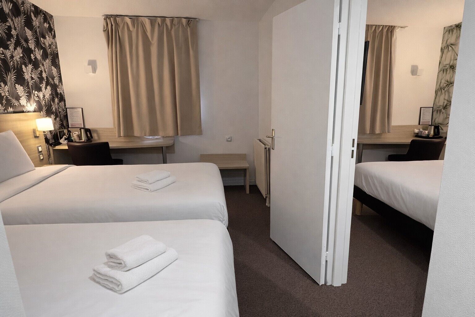 A hotel room with two beds, a desk, a chair, a small table, a radiator, and a mirror.