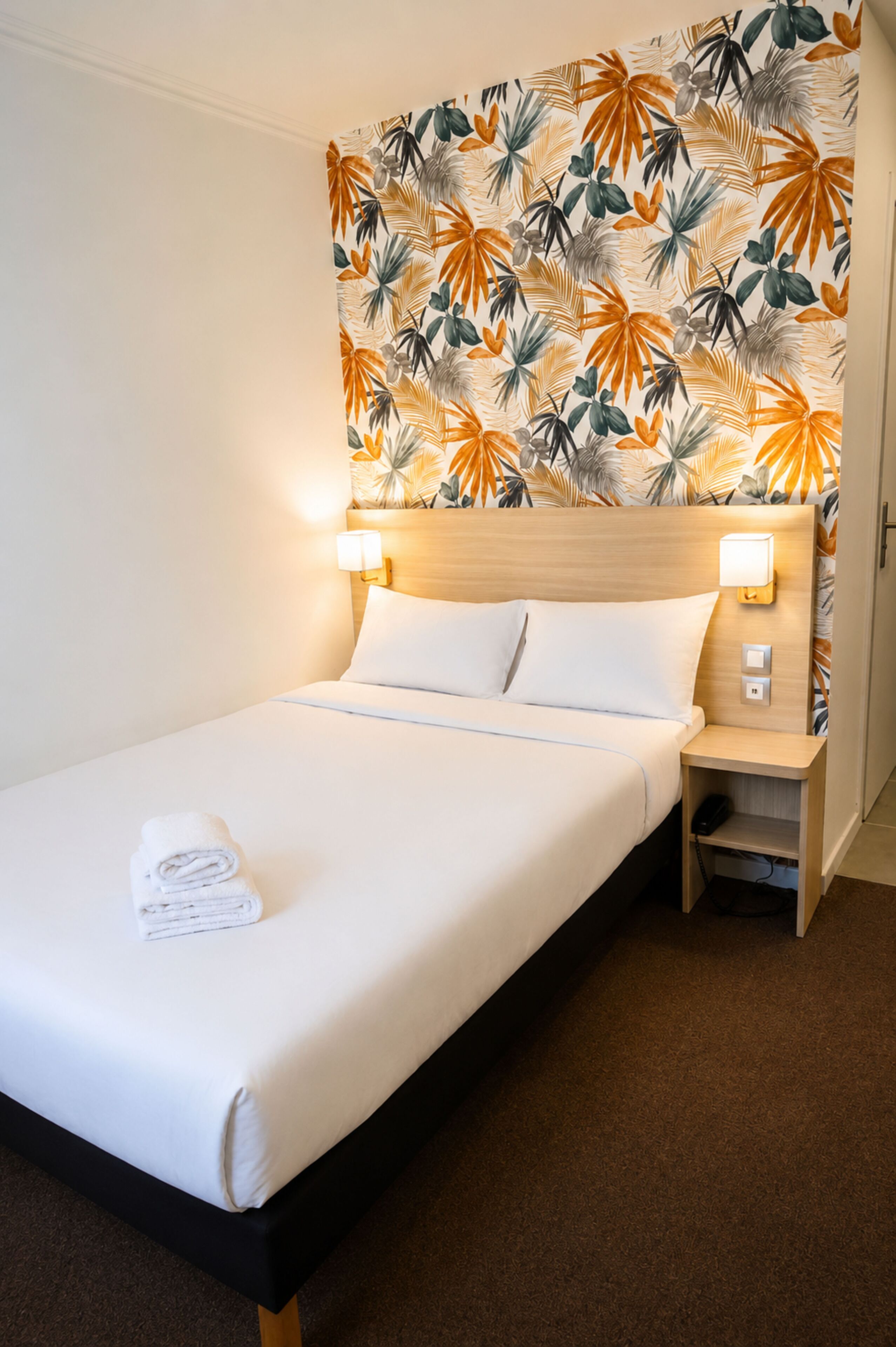 A hotel room with a bed, two bedside lamps, a nightstand, and a wall with a tropical leaf pattern.