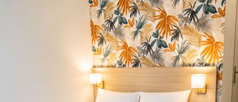 Una habitación de hotel con una cama, dos lámparas de noche, una mesita de noche y una pared con un estampado de hojas tropicales.