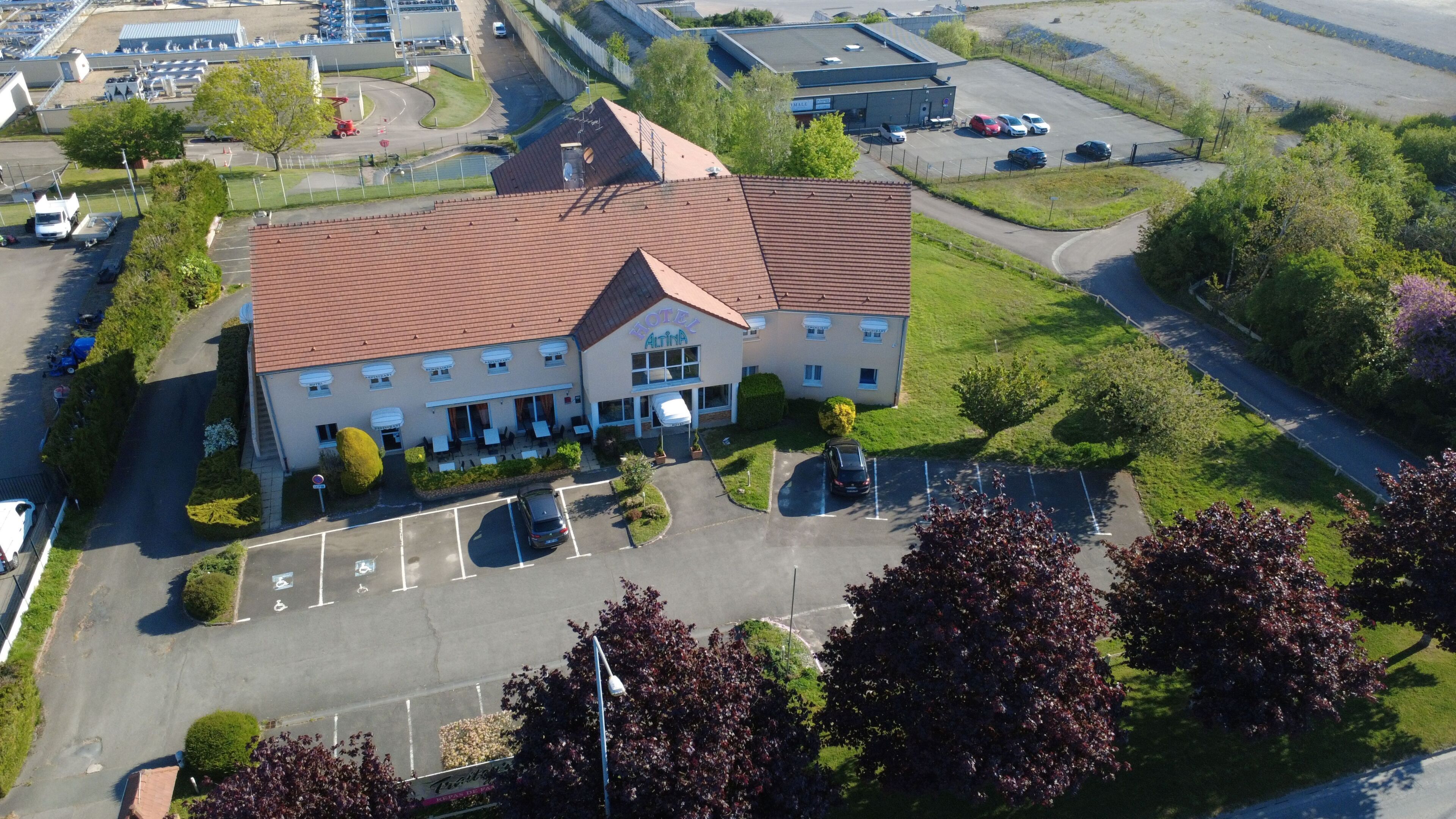 Vue aérienne d’un hôtel avec un toit recouvert de tuiles rouges, un parking et des espaces verts environnants.