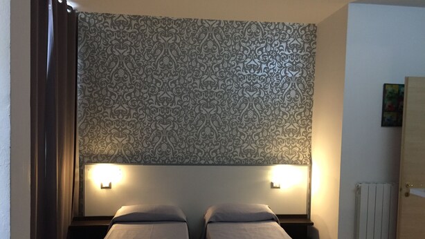 A room with two single beds, a patterned wallpaper, two bedside lamps, a radiator, and a door. - Scacco Matto (Milan)