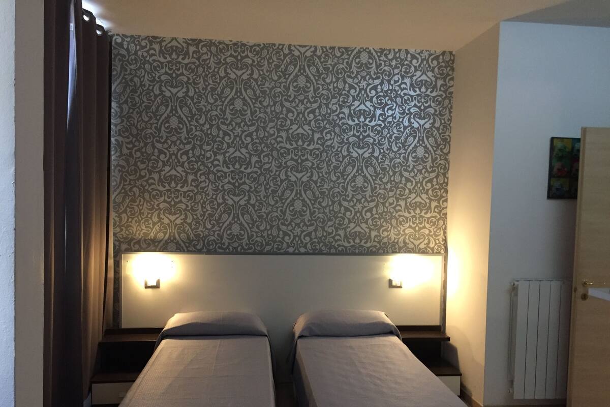 A room with two single beds, a patterned wallpaper, two bedside lamps, a radiator, and a door.