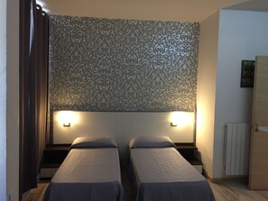 Una habitación con dos camas individuales, papel tapiz estampado, dos lámparas de noche, un radiador y una puerta.