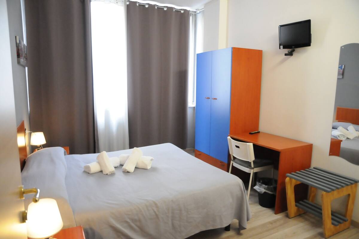 A hotel room with a bed, a desk, a chair, a TV, and a window with curtains.