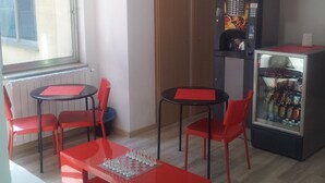 A small kitchenette with a microwave, refrigerator, and coffee machine. There are two red tables with chairs, and a window providing natural light. - Scacco Matto (Milan)