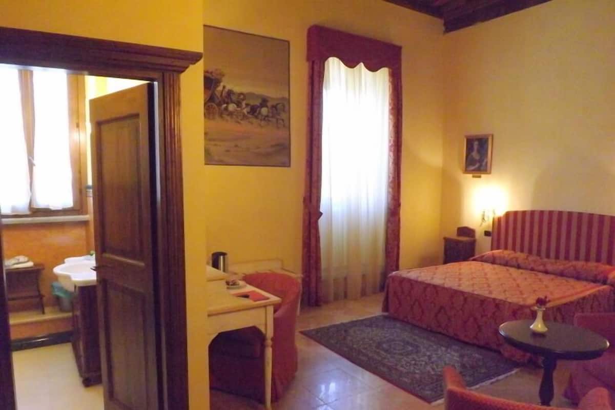 A hotel room with a bed, a desk, a chair, a small table, and a painting on the wall.