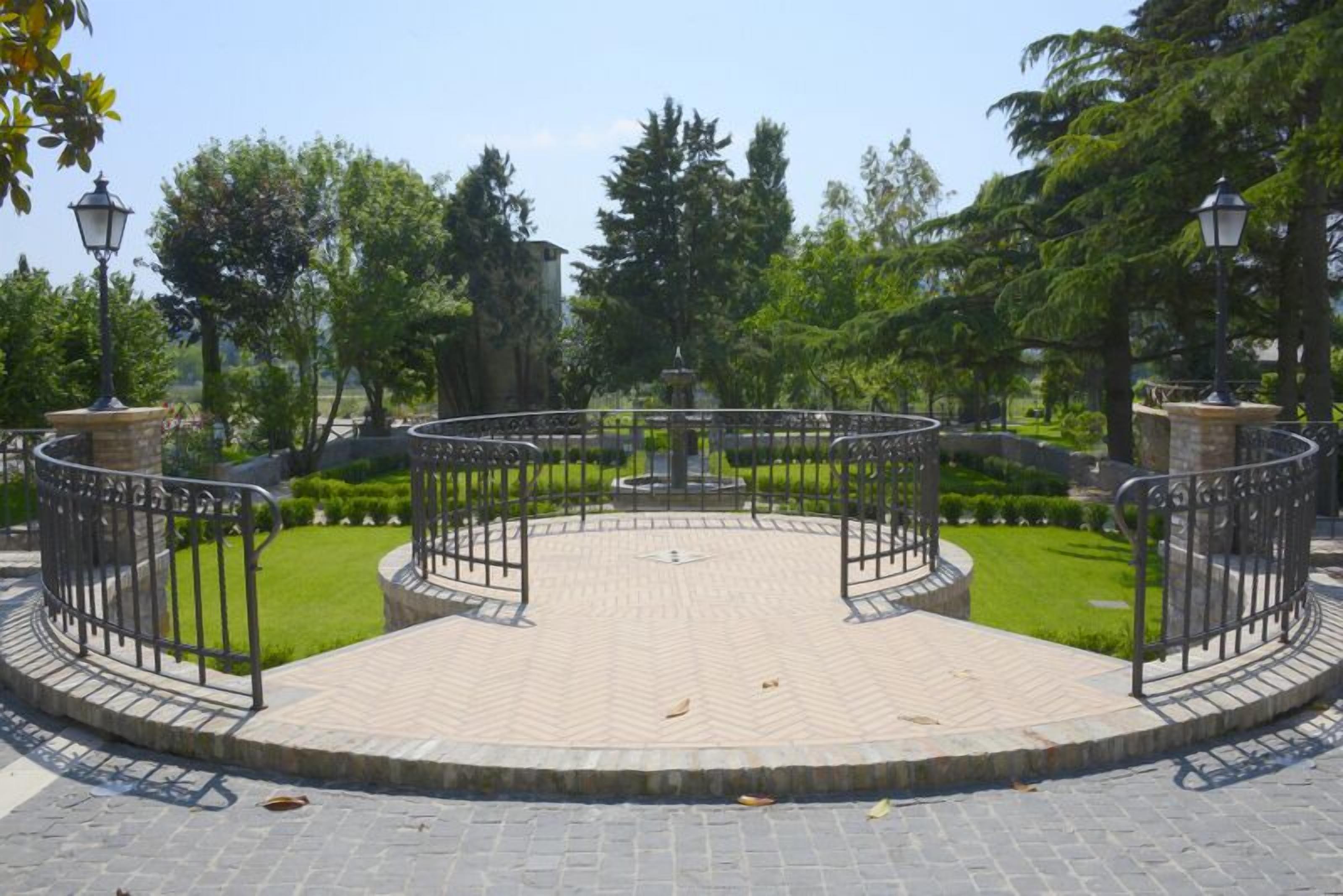 A circular paved area with a central fountain, surrounded by a metal fence and stone pillars.