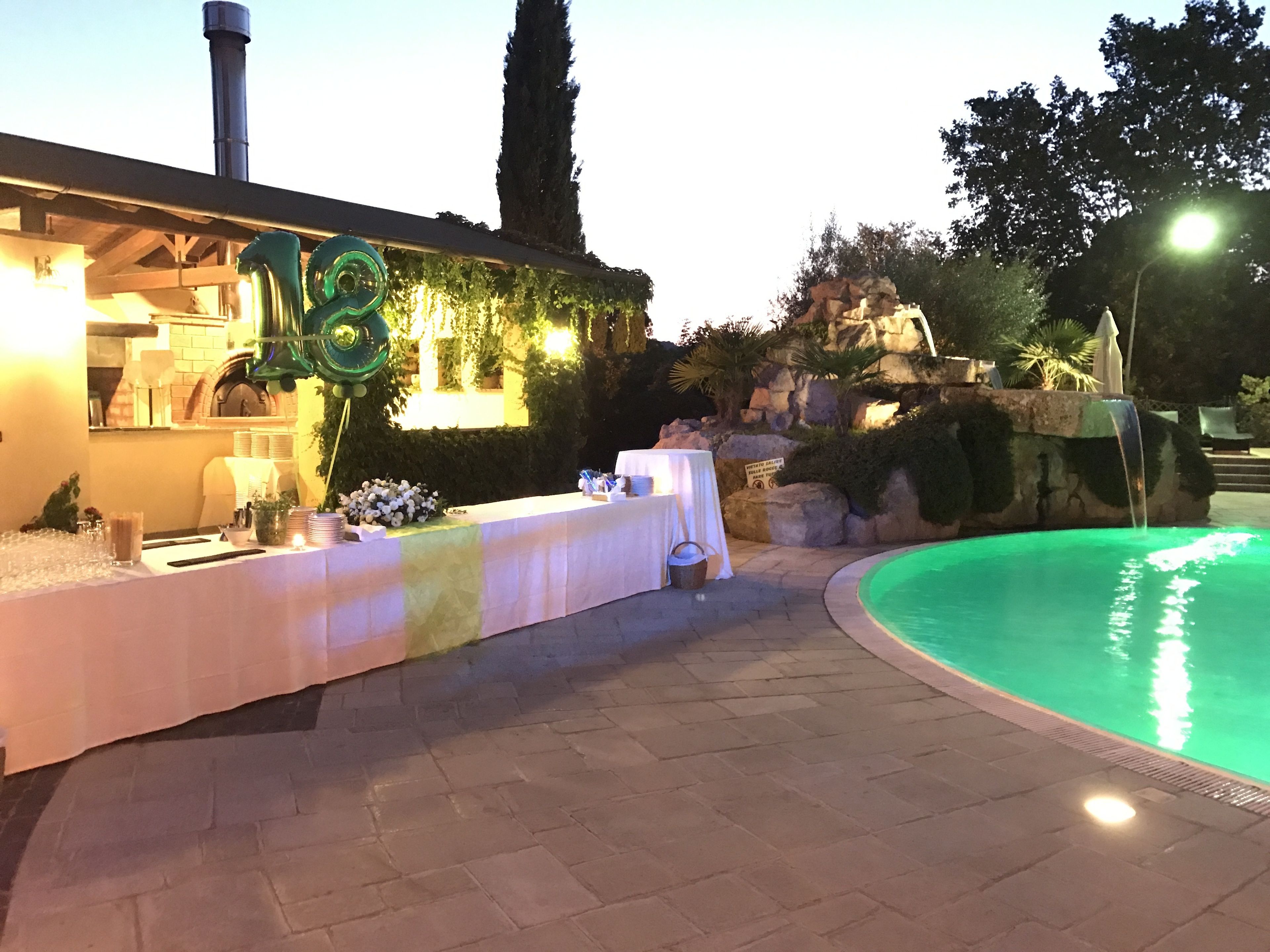 An outdoor pool area with a table set up for a reception or event, surrounded by greenery and a stone wall.