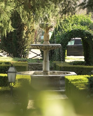 Een stenen fontein waarvan water uit de bovenkant spuit, omgeven door goed onderhouden hagen en bomen.