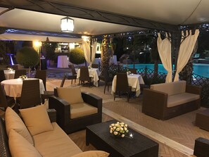Een terras buiten met rieten meubels, tafels en uitzicht op een zwembad.