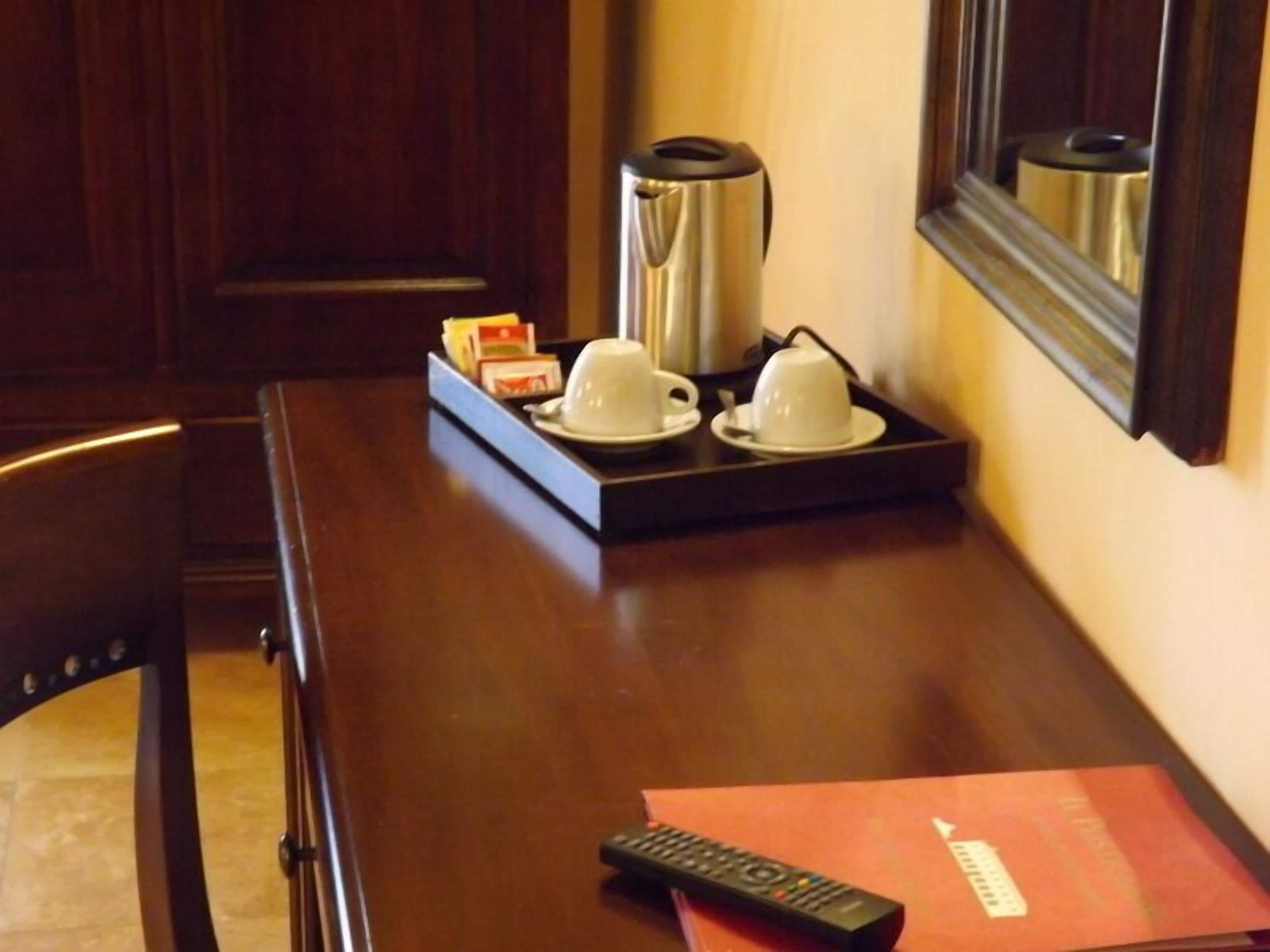 A hotel room with a wooden table, two white cups on a tray, a remote control, and a red booklet.