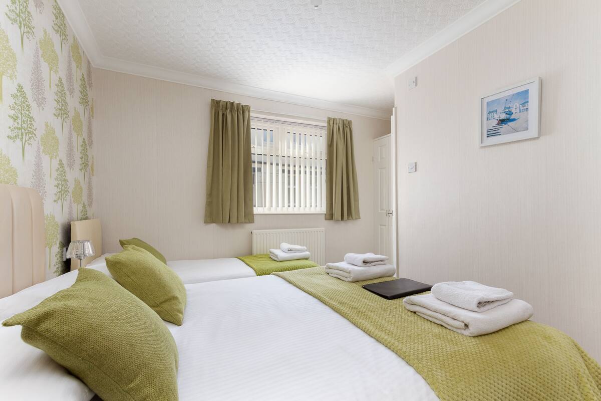 A hotel room with two beds, green pillows, white bedding, a window with curtains, a radiator, a framed picture on the wall, and a bedside table with a lamp and a book.