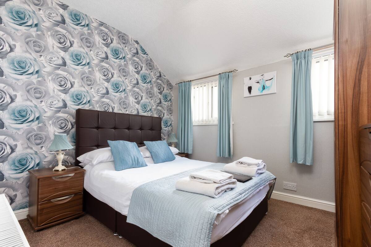 A bedroom with a large bed, a nightstand, a wall with a floral pattern, and light blue curtains.
