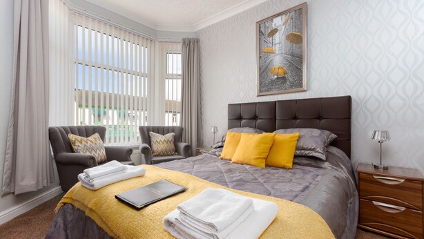 A modern hotel room with a large bed, a chair, a bedside table, and a window with curtains. - The Almeria (Blackpool)